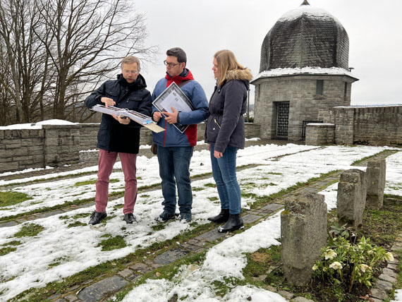Das Bild zeigt drei Personen auf dem Friedhof beim Betrachten von Aufzeichnungen