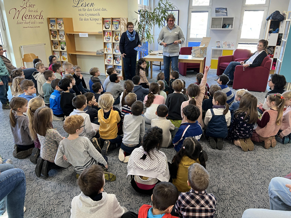 Das Bild zeigt eine Gruppe von Schülerinnen und Schülern bei einer Leseaktion mit zwei Bibliothek-Mitarbeiterinnen der Stadtbibliothek 