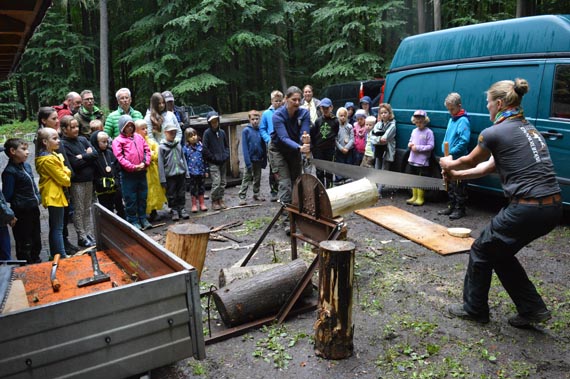Das Bild zeigt eine große Gruppe von Kindern im Wald, die zwei Erwachsenen beim Timber--Sport zuschauen.