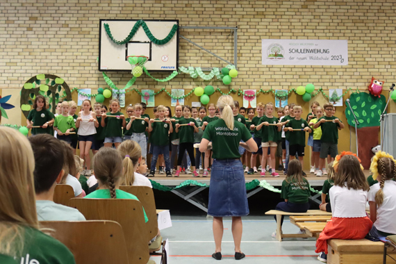 Das Bild zeigt eine Gruppe von Grundschulkindern beim musikalischen Vortrag auf einer Bühne und einige erwachsene Zusehende,