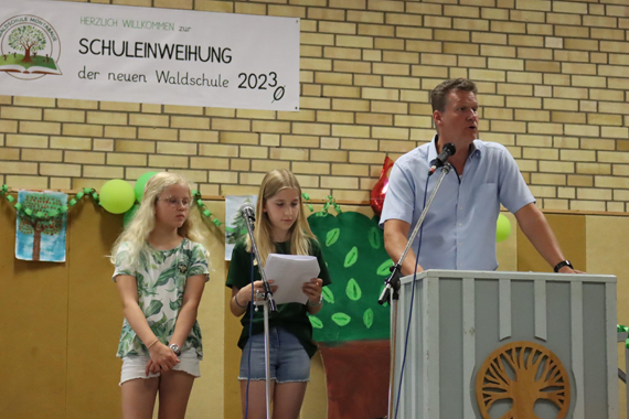 Das Bild zeigt einen Erwachsenen am Rednerpult in einer Turnhalle und zwei Mädchen im Grundschulalter