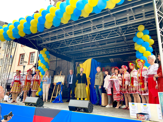 Das Bild zeigt eine Personen-Gruppe auf einer Bühne, rechts und links flankiert von Akteuren einer Folkloregruppe