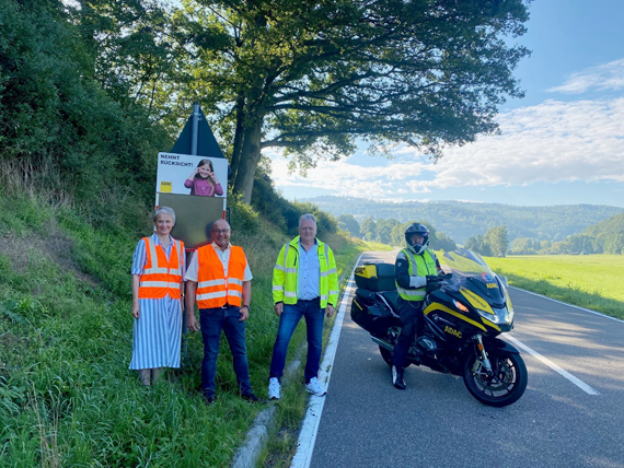 Das Bild zeigt vier Erwachsene - mit Warnwesten bekleidet  (davon ein Motorradfahrer) - auf einer Landstraße 