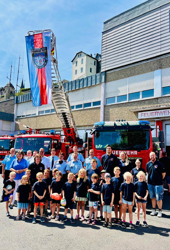 Außenaufnahme einer großen Gruppe Erwachsener und Kinder und Feuerwehrfahrzeuge vor einem Feuerwehrgebäude.