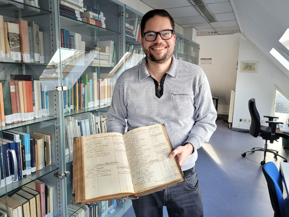 Das Bild zeigt einen Mann vor einem Bücherregal, der ein offenes Buch präsentiert