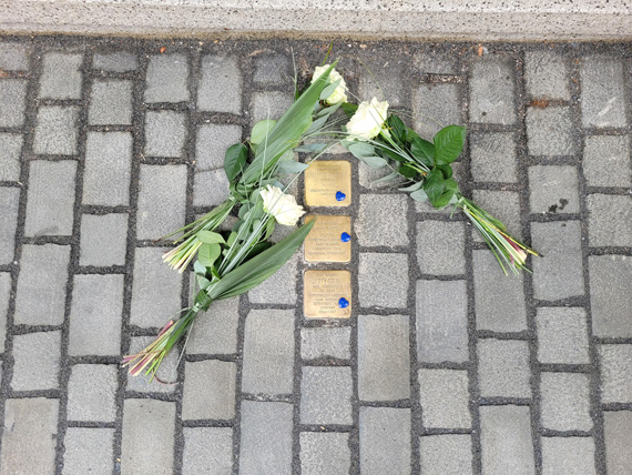 Das Bild zeigt drei kleine Messingtafeln, die ins Straßenpflaster integriert wurden und darauf platziert drei weiße Rosen.