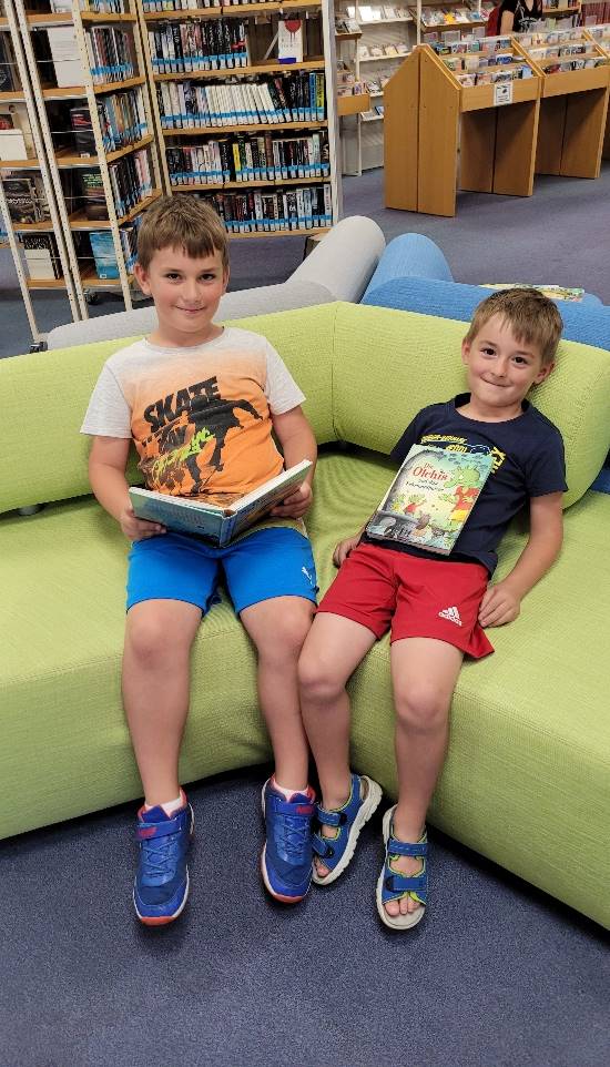 Das Bild zeigt zwei Kinder auf einer Couch sitzend mit einem Buch in der Hand