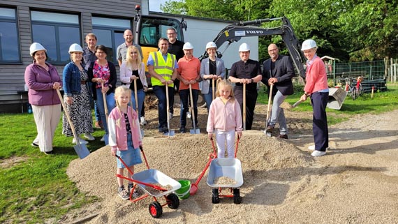 Das Bild zeigt eine Gruppe von Erwachsenen und Kindern mit Schaufeln und Schubkarren auf einer Baustelle