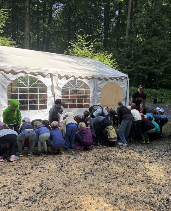 Das Bild zeigt eine Gruppe von Kindern und Erwachsenen beim Versuch, gemeinsam einen Baumstamm anzuheben